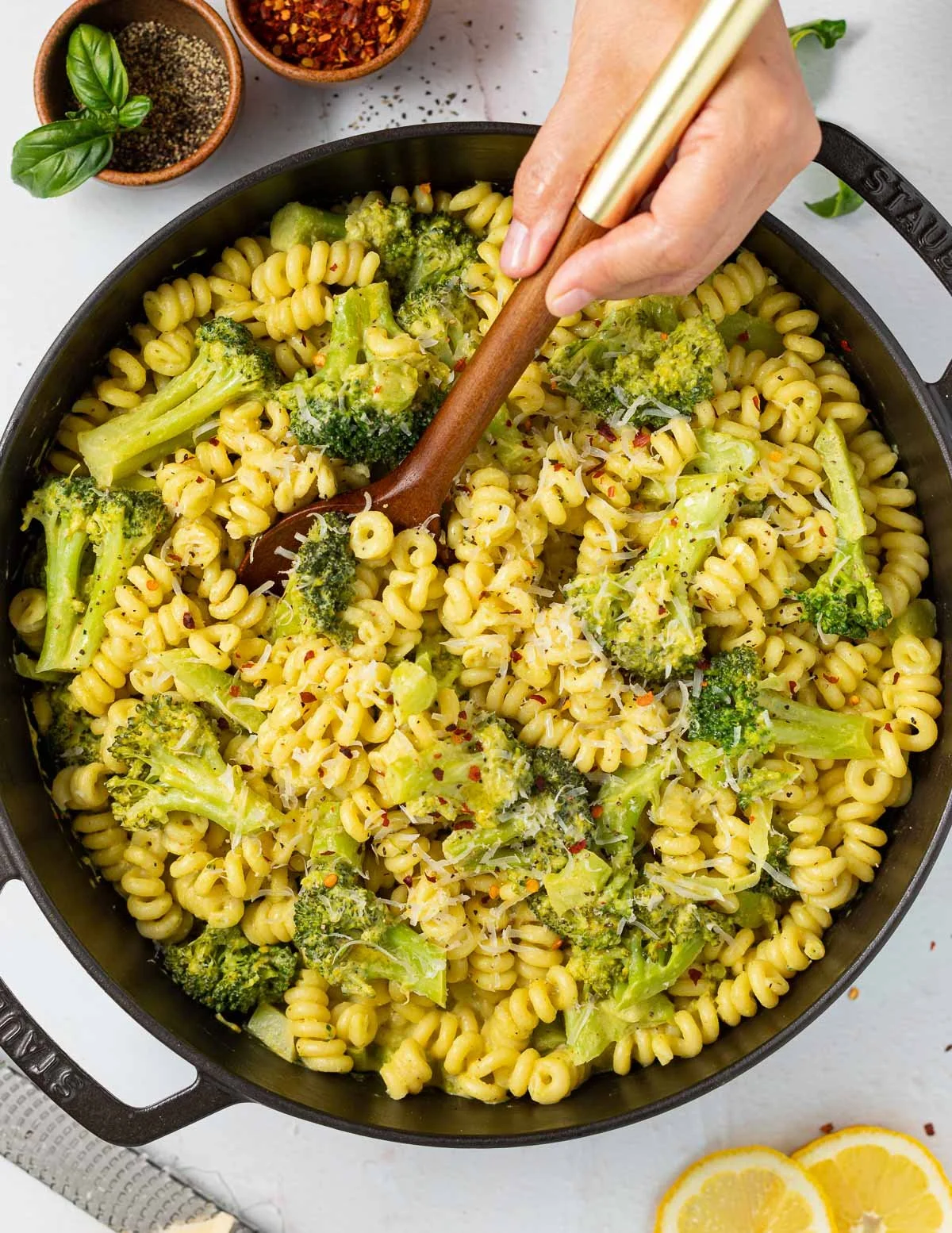 A skillet filled with cooked pasta and broccoli, being stirred with a wooden spoon, with bowls of spices and lemon slices nearby.