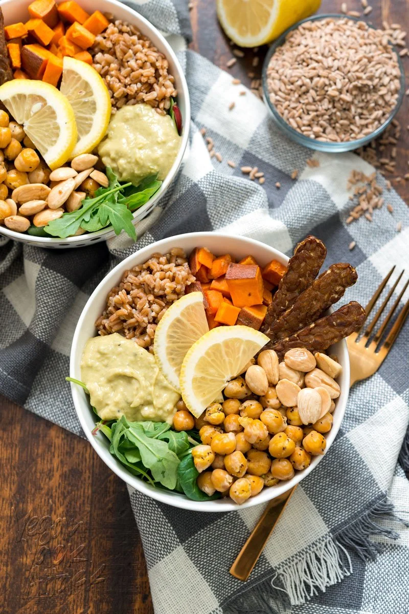Two bowls of vegan poke bowls with chickpeas, sweet potatoes, avocado, lemon slices, and crispy tempeh served on a checkered napkin with a fork.