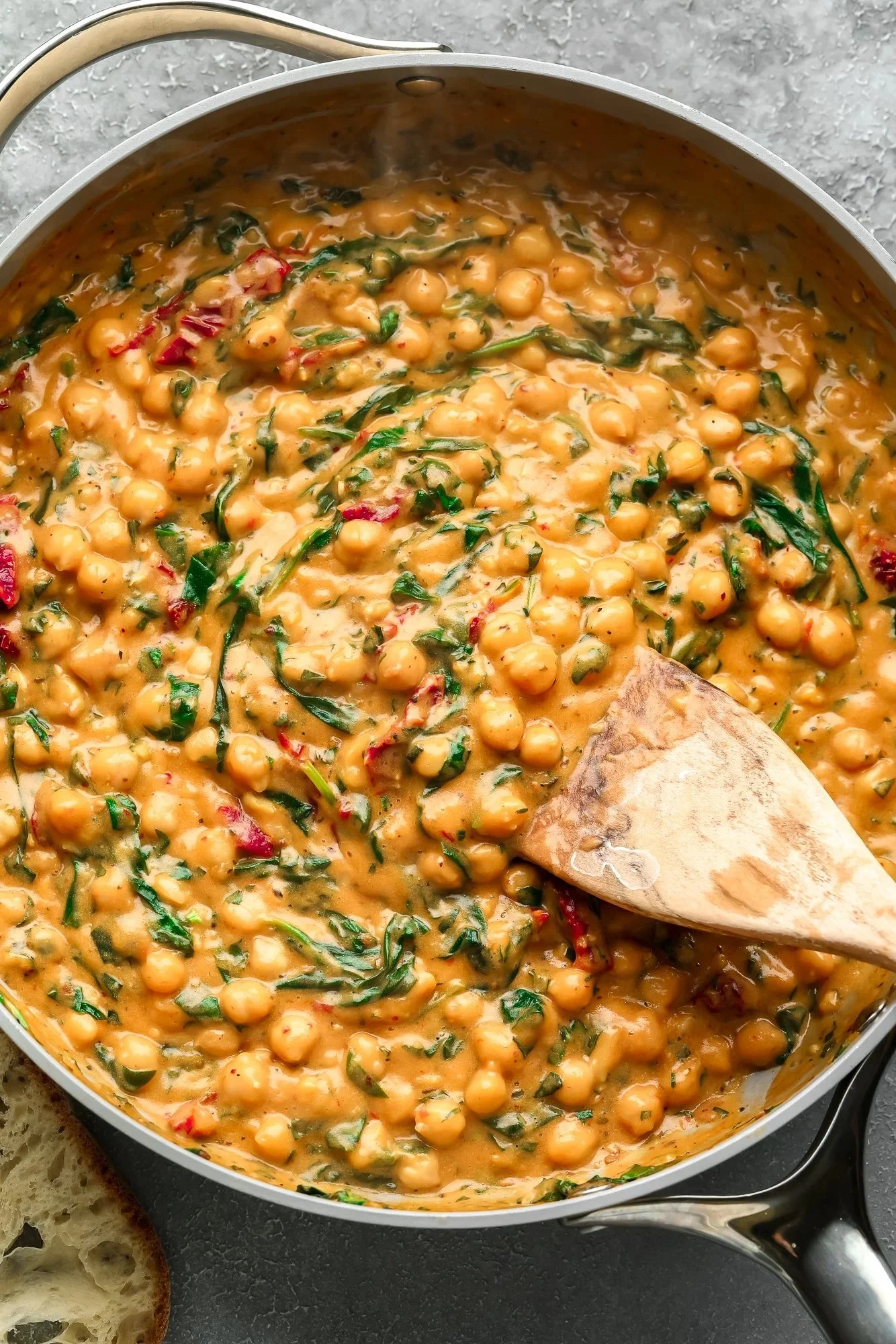 A pan of chickpea and spinach curry being cooked, with a wooden spatula resting in the pan, on a dark countertop, with a piece of bread partially visible in the corner.