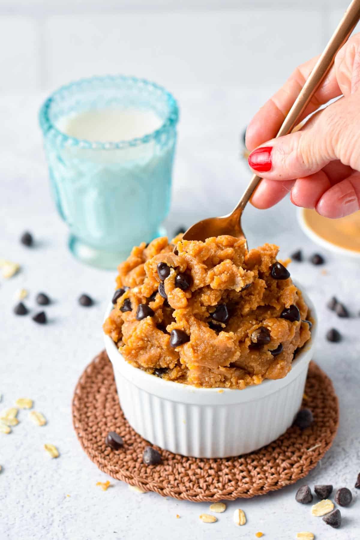 Close-up of a white ramekin filled with peanut butter cookie dough with chocolate chips, on a round woven coaster. A hand with red nail polish is holding a spoon over the dough. There is a glass of milk in the background and scattered chocolate chips and oats on the surface.