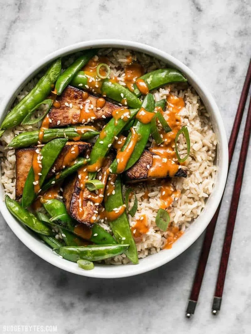 Bowl of cooked rice topped with tofu, sliced green onions, sugar snap peas, and a drizzle of orange sauce, with chopsticks placed beside the bowl on a marble surface.