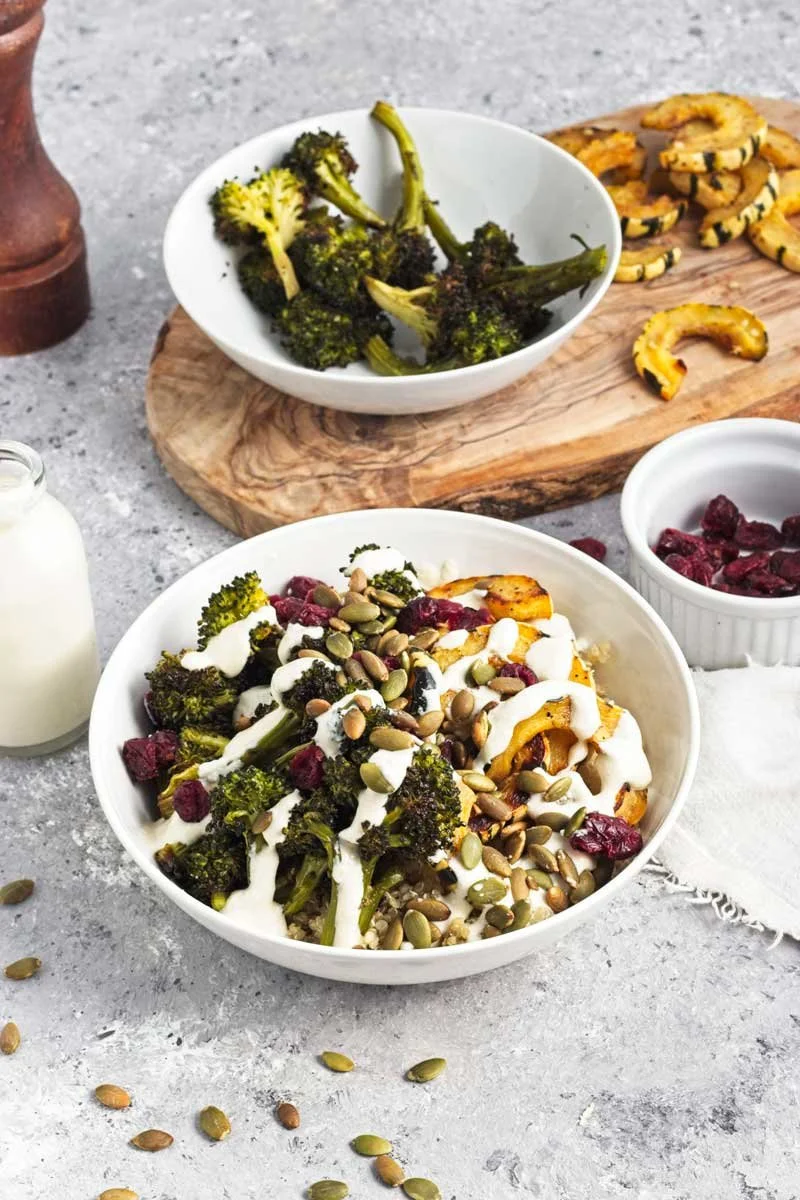 A bowl of roasted broccoli topped with a white sauce and pumpkin seeds, alongside a small bowl of cranberries, with roasted squash on a wooden board and a milk bottle in the background.
