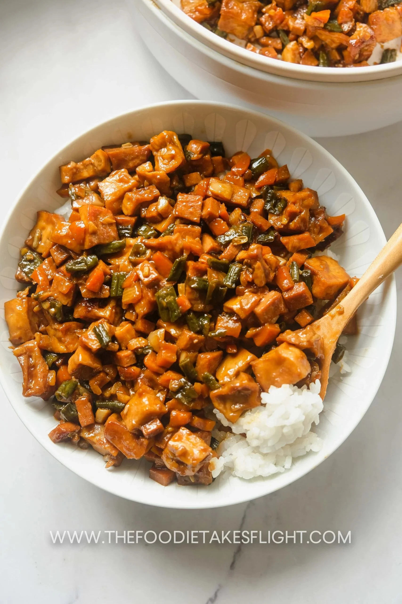 White bowl of rice topped with stir-fried diced chicken, carrots, and green beans in sauce, with another similar bowl in the background.