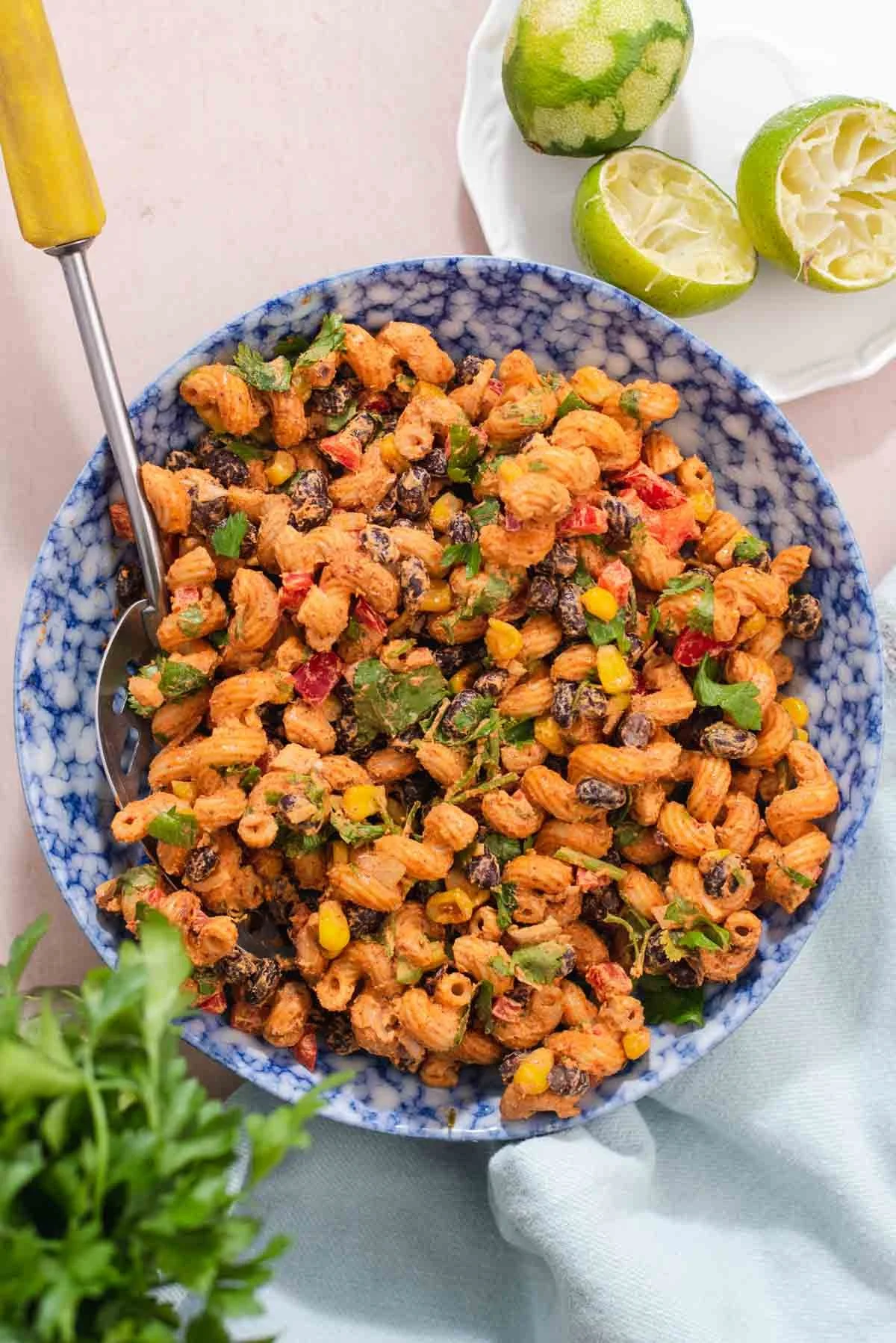 A large bowl of vegan taco salad pasta with corn, black beans, tomatoes, cilantro, and greens, with lime halves on a plate nearby.