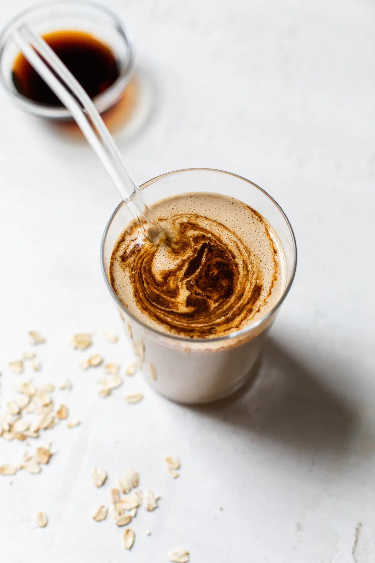 A glass of coffee with vegan milk being poured into it, creating a swirl. There is a small bowl of dark coffee in the background and some scattered oats on a white surface.