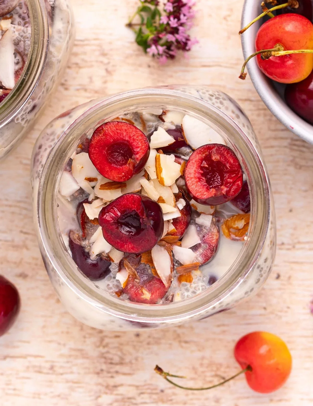 A glass jar filled with cherries, coconut flakes, and chopped almonds on a wooden surface, with a bowl of cherries and a small bunch of flowers nearby.