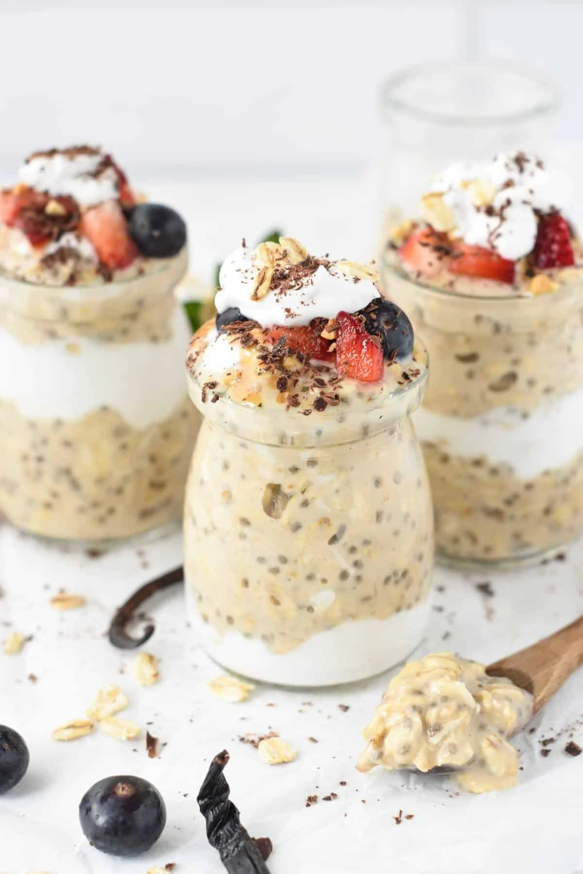 Creamy overnight oats in small glass jars topped with whipped cream, strawberries, blueberries, chopped nuts, and chocolate shavings, with scattered blueberries and oats around on a white surface.