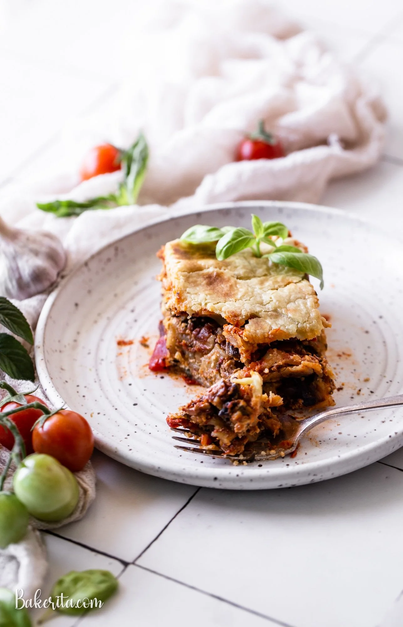 A slice of vegan lasagna garnished with a basil leaf on a speckled white plate, surrounded by cherry tomatoes and green olives on a white tiled surface with a white cloth and garlic in the background.