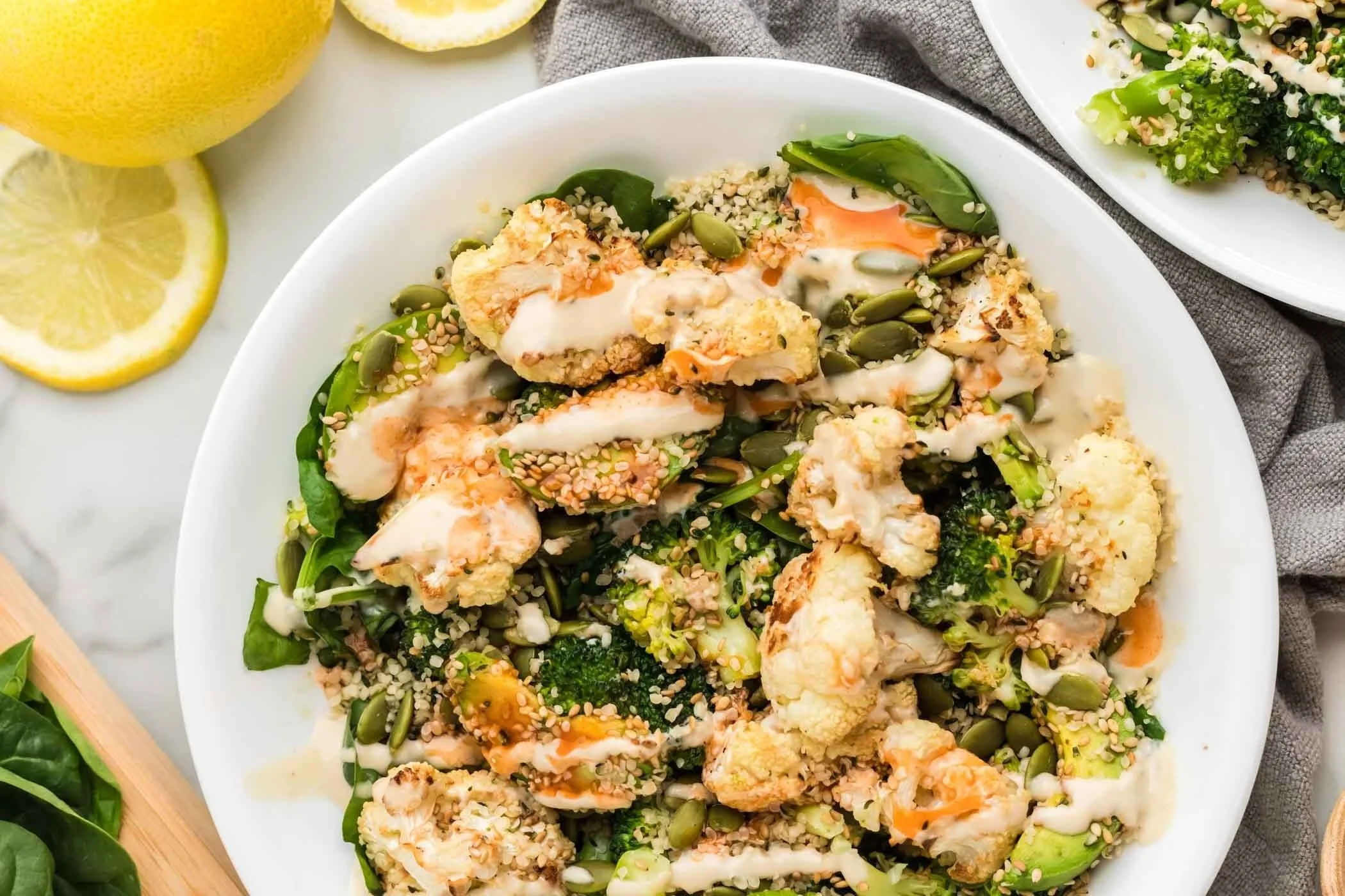 A white bowl filled with a salad containing roasted cauliflower, spinach, broccoli, sunflower seeds, pumpkin seeds, and a creamy dressing. Lemon wedges are visible in the background.