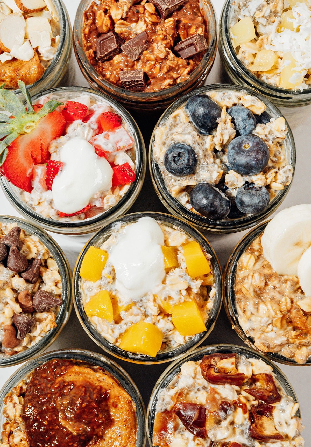 Eight small jars filled with various flavors of overnight oats topped with fruits and toppings, including strawberries, blueberries, mango, chocolate chunks, shredded coconut, and nuts.