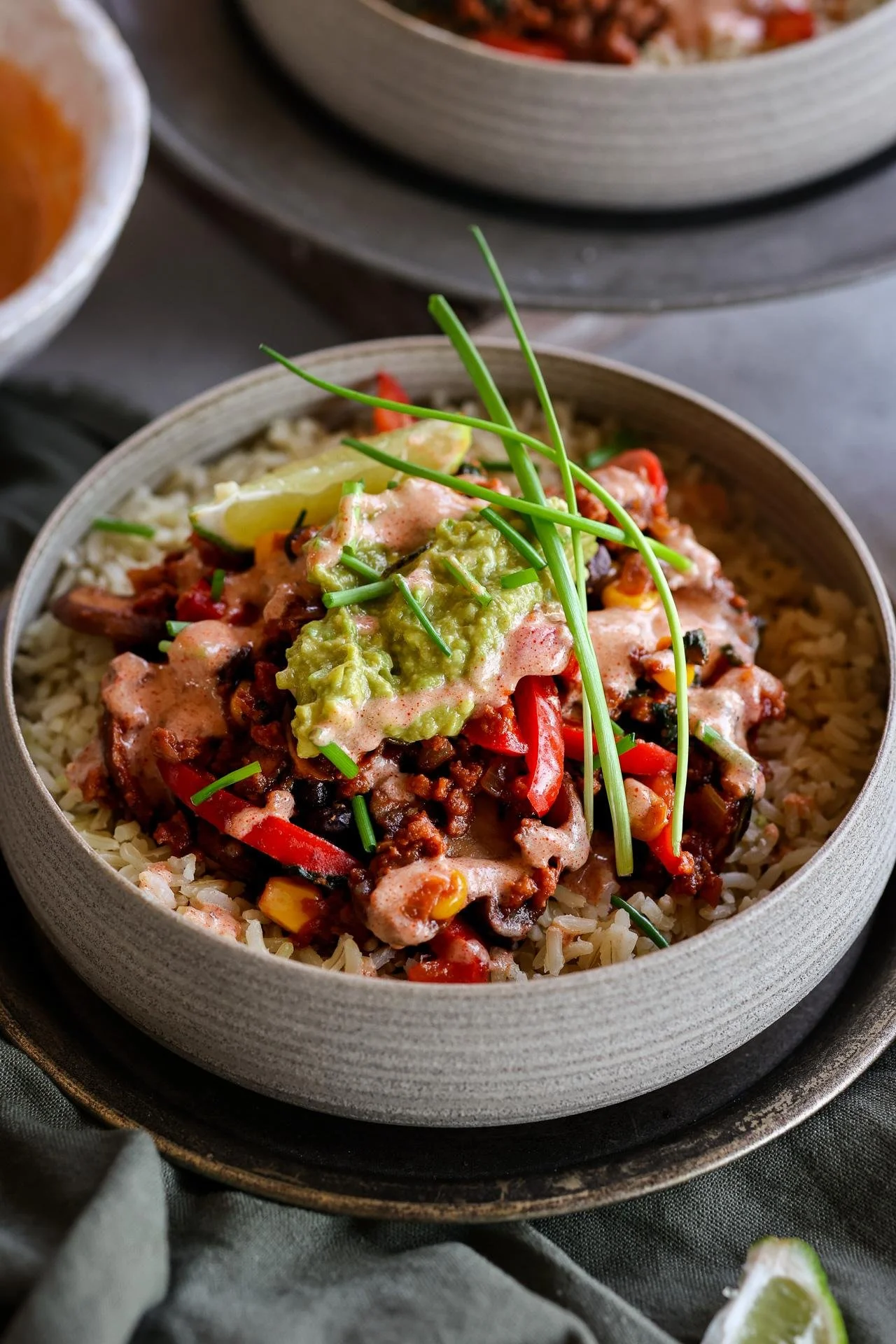 A bowl of seasoned rice topped with chili, guacamole, chopped chives, and lime wedges, garnished with green chive stems.