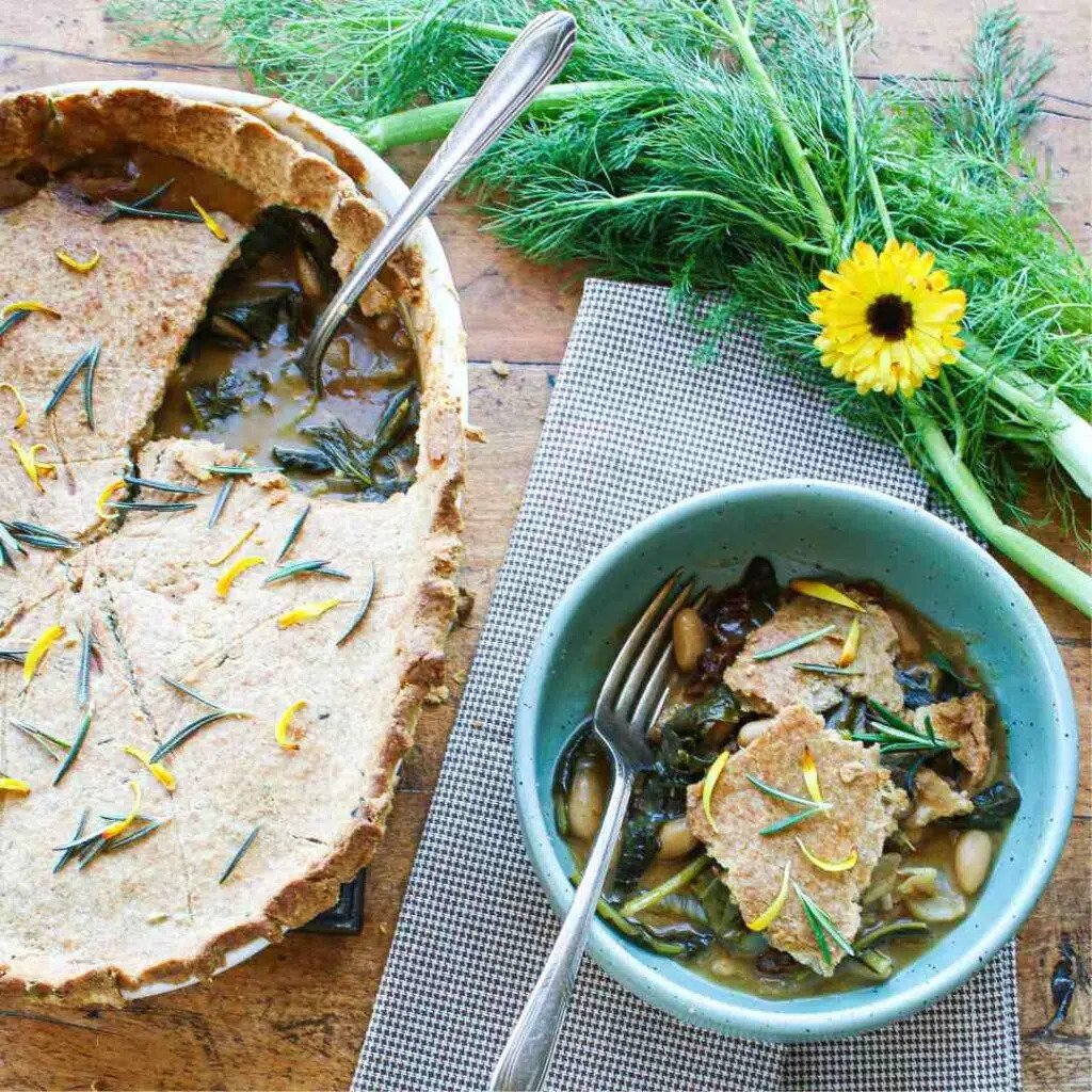 A rustic table setting with a vegetable pot pie with a slice taken out, a bowl with a serving of the pie, and fresh herbs and a yellow flower for decoration.