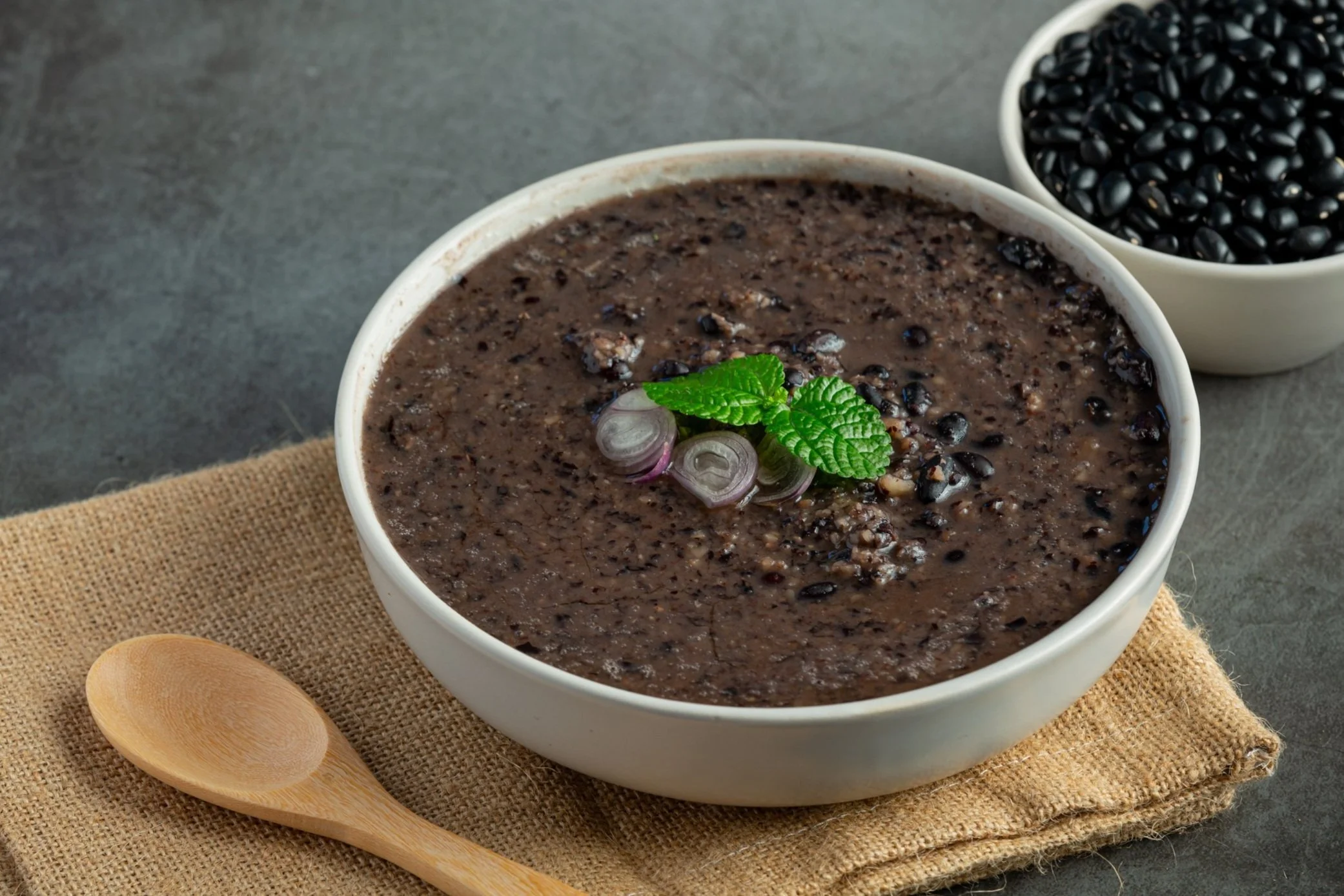 A bowl of black bean soup garnished with sliced onions and mint leaves, with a small bowl of black beans nearby on a gray surface.