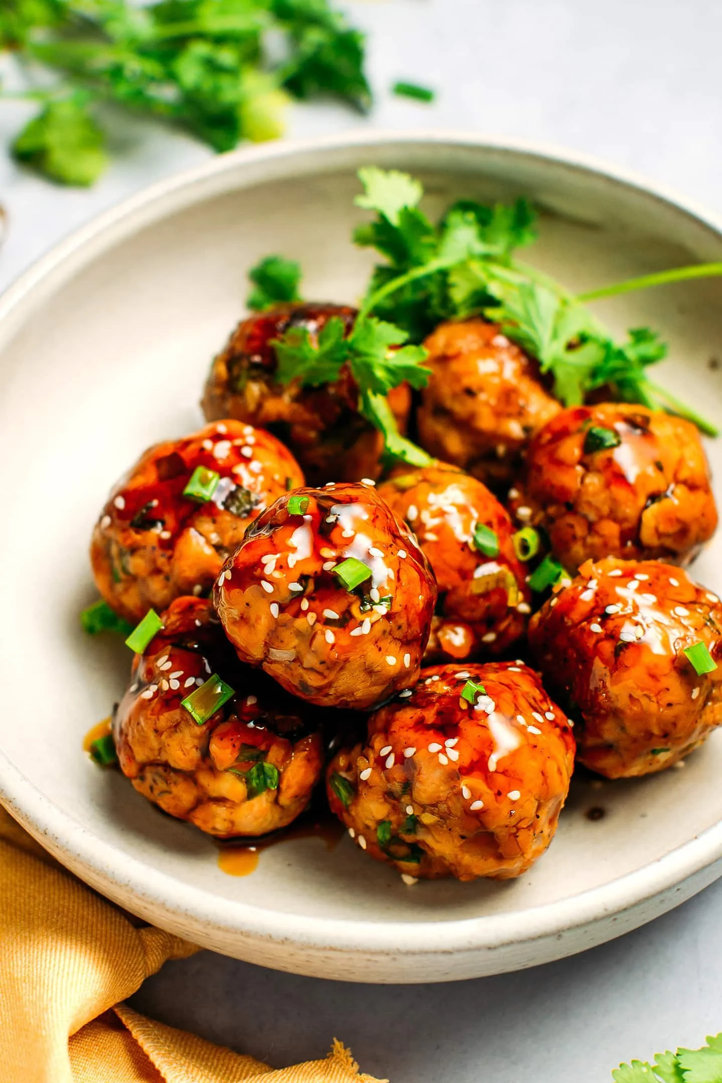 A bowl of vegan Asian-style meatballs garnished with chopped green onions and sesame seeds, topped with a drizzle of sauce, served with cilantro.