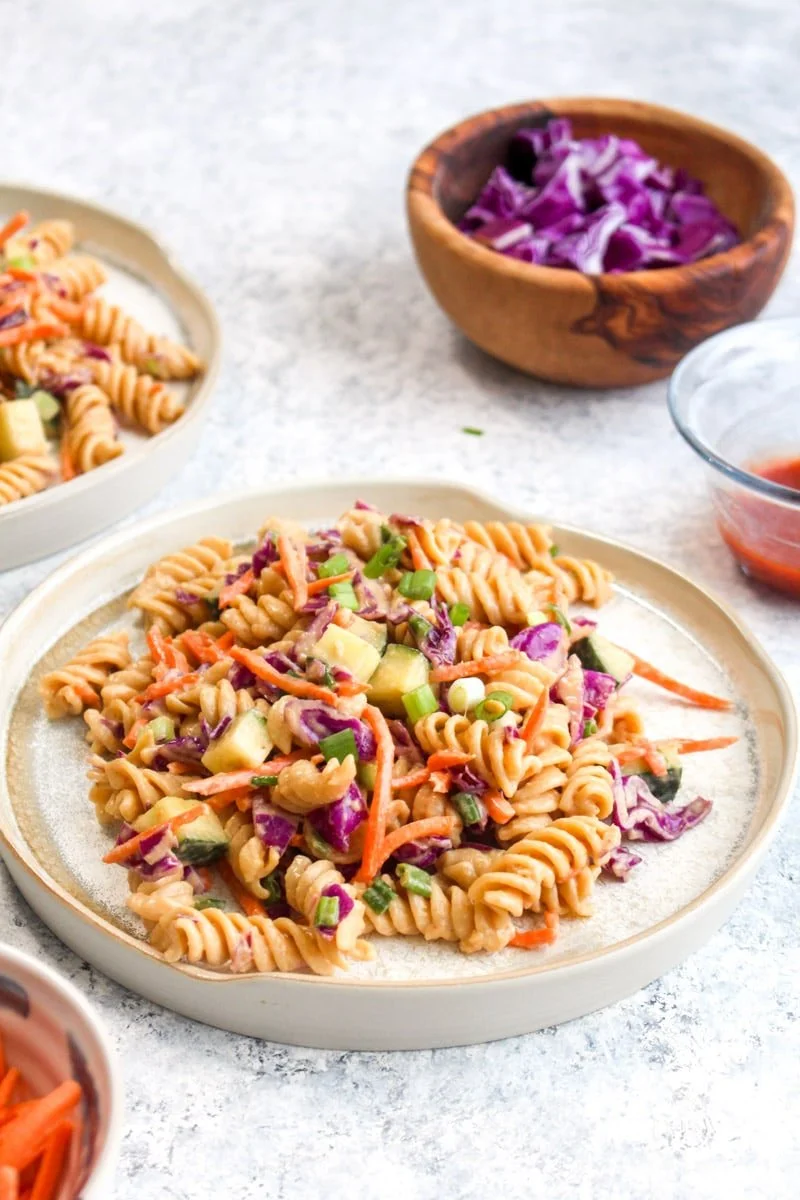 A plate of cold pasta salad with rotini pasta, cucumber, shredded carrots, purple cabbage, and chopped green onions, garnished with chopped green herbs, with bowls of purple cabbage and dressings in the background.