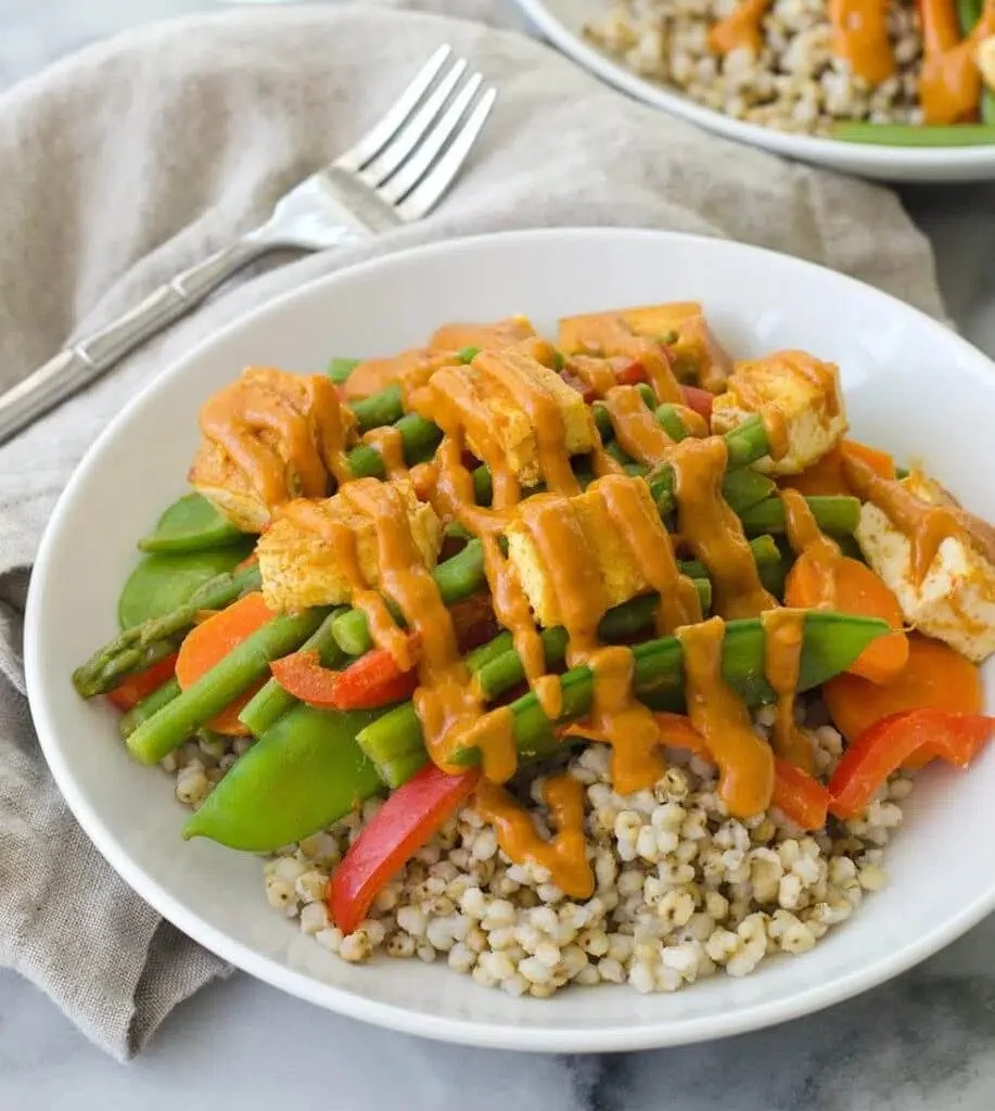 Bowl of cooked barley topped with sautéed vegetables, tofu, and drizzled with sauce
