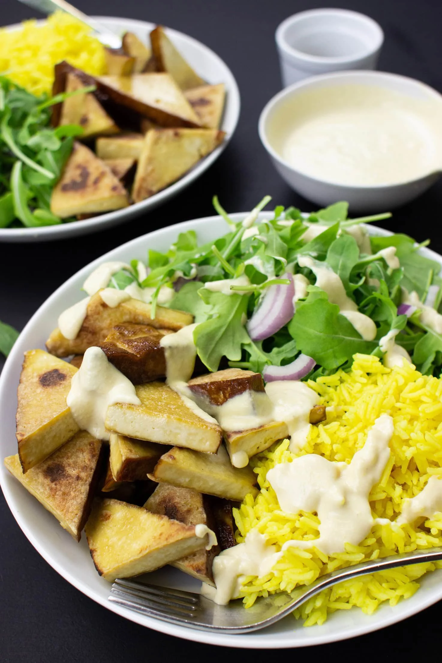 A plate of yellow rice, roasted potatoes, and salad with greens, chopped onions, and dressing, with additional roasted potatoes, salad, and sauces in the background.