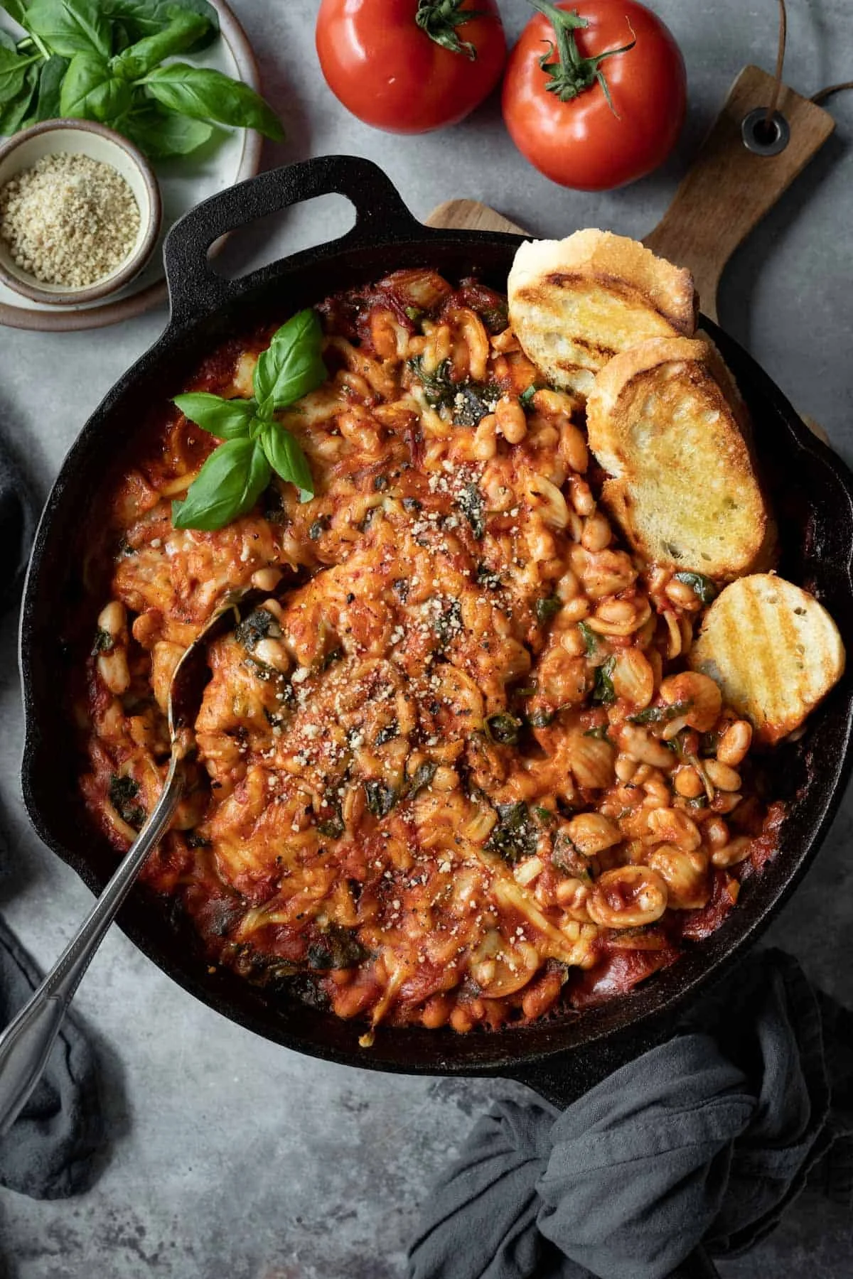 A skillet of baked beans topped with grated cheese, basil, and grilled bread slices on the side, with tomatoes and herbs in the background.