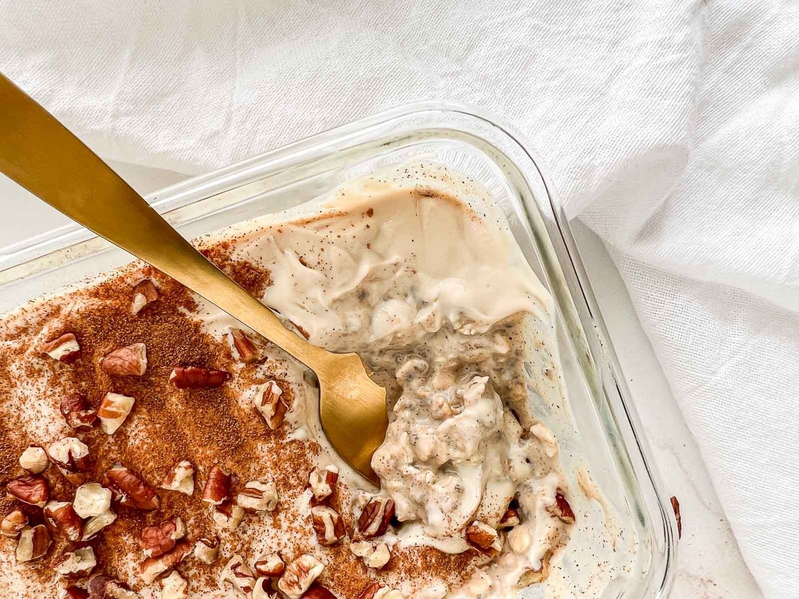 A glass baking dish with a layered dessert topped with chopped pecans and cinnamon, a gold spoon resting inside, placed on a white cloth surface.