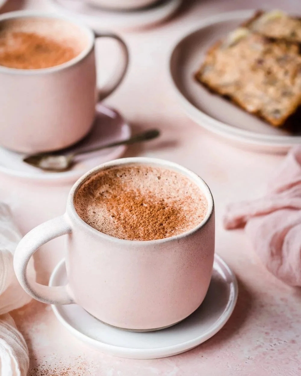 A close-up of a pink coffee mug filled with frothy hot chocolate topped with cinnamon, on a pink table with a matching saucer. In the background, there is a second mug and a plate with a scone.