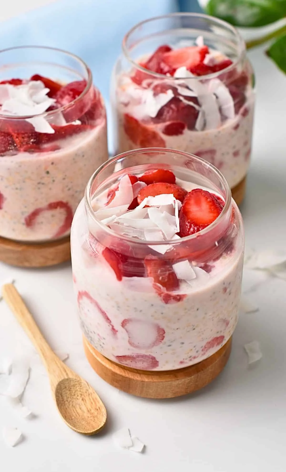 Three glasses of strawberry chia pudding topped with fresh strawberries and coconut flakes, placed on wooden coasters with a small wooden spoon and scattered coconut flakes nearby.
