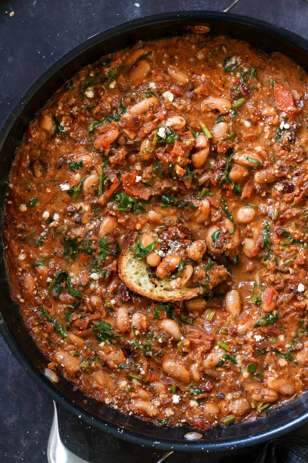 Hearty white bean soup with vegetables and herbs in a black skillet, garnished with a slice of toasted bread and crumbled cheese.