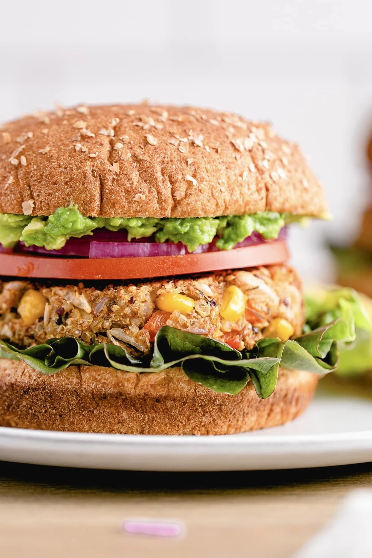 Close-up of a veggie burger on a white plate, featuring a whole wheat bun, avocado, tomato, red onion, lettuce, and a veggie patty with corn and vegetables.