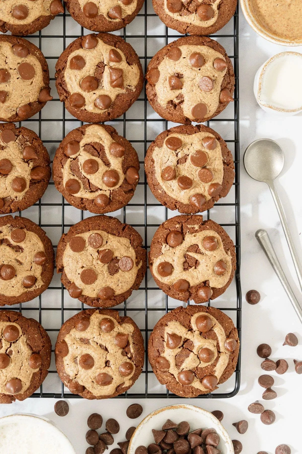 A batch of vegan chocolate chip cookies cooling on a wire rack, with scattered chocolate chips and bowls of milk around them.