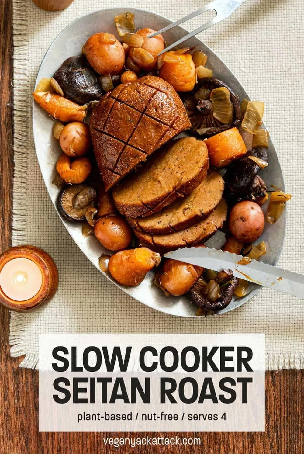 A platter of sliced seitan roast surrounded by roasted vegetables including carrots, potatoes, mushrooms, and onions, with a small lit candle beside it. There is a knife on the platter for serving.