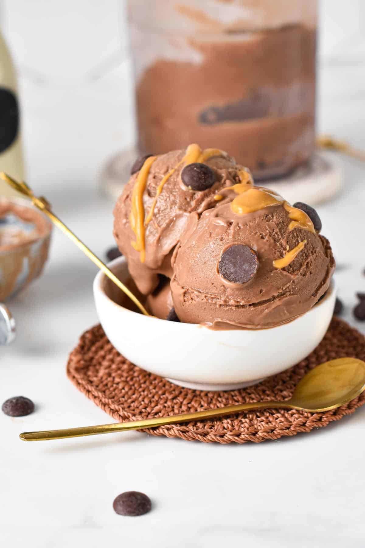 Three scoops of chocolate ice cream with chocolate chips and caramel drizzle in a white bowl on a woven placemat with a gold spoon beside it.