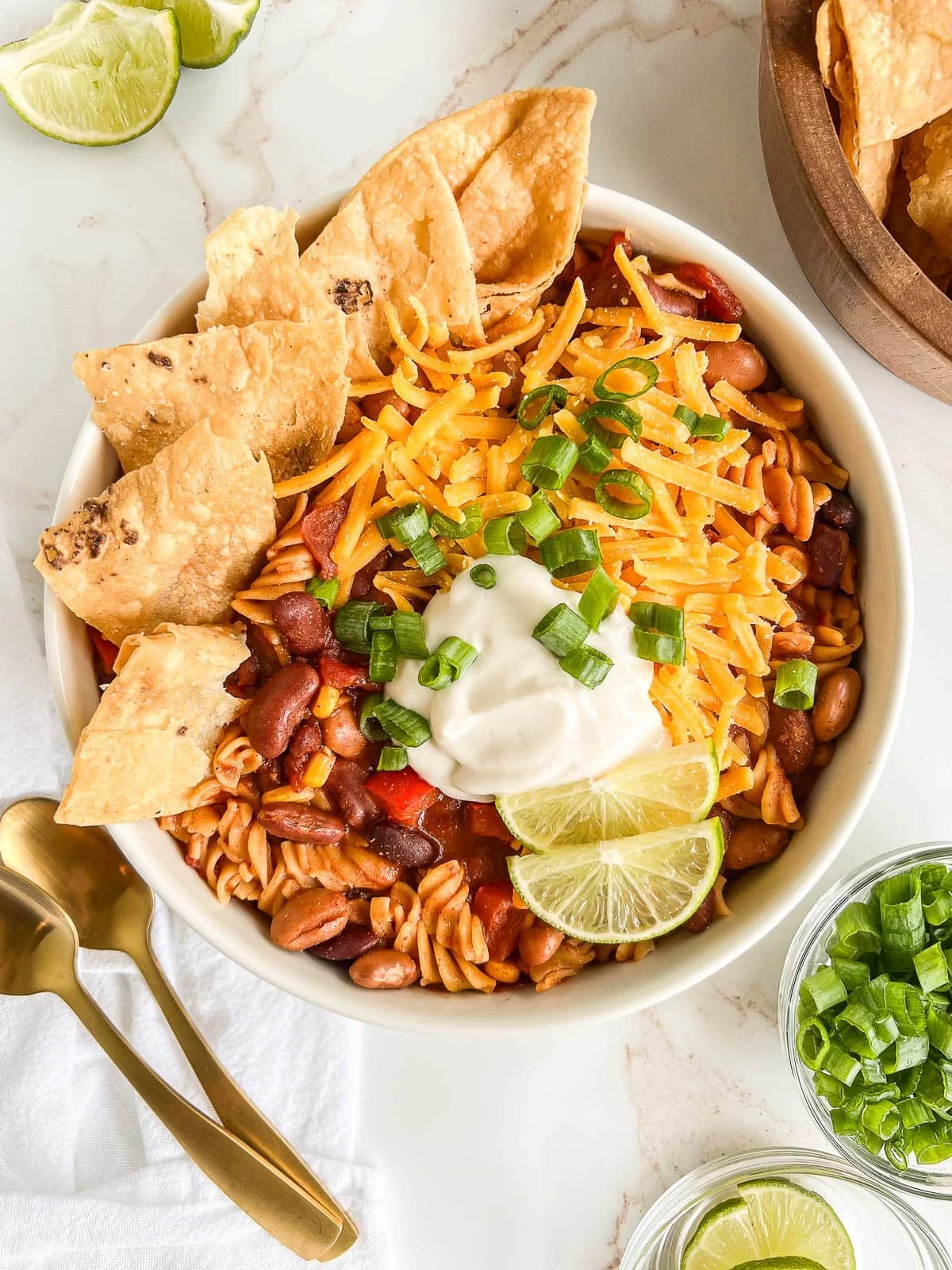 Bowl of vegan southwest pasta salad with black beans, shredded cheese, sour cream, green onions, and tortilla chips, garnished with lime wedges.