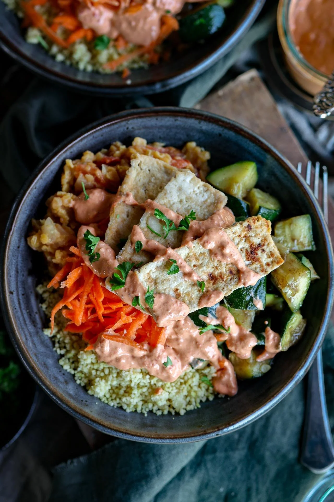 A bowl containing grilled paneer, mixed vegetables including zucchini, shredded carrots, and couscous, with a drizzle of creamy pink sauce and fresh herbs on top.
