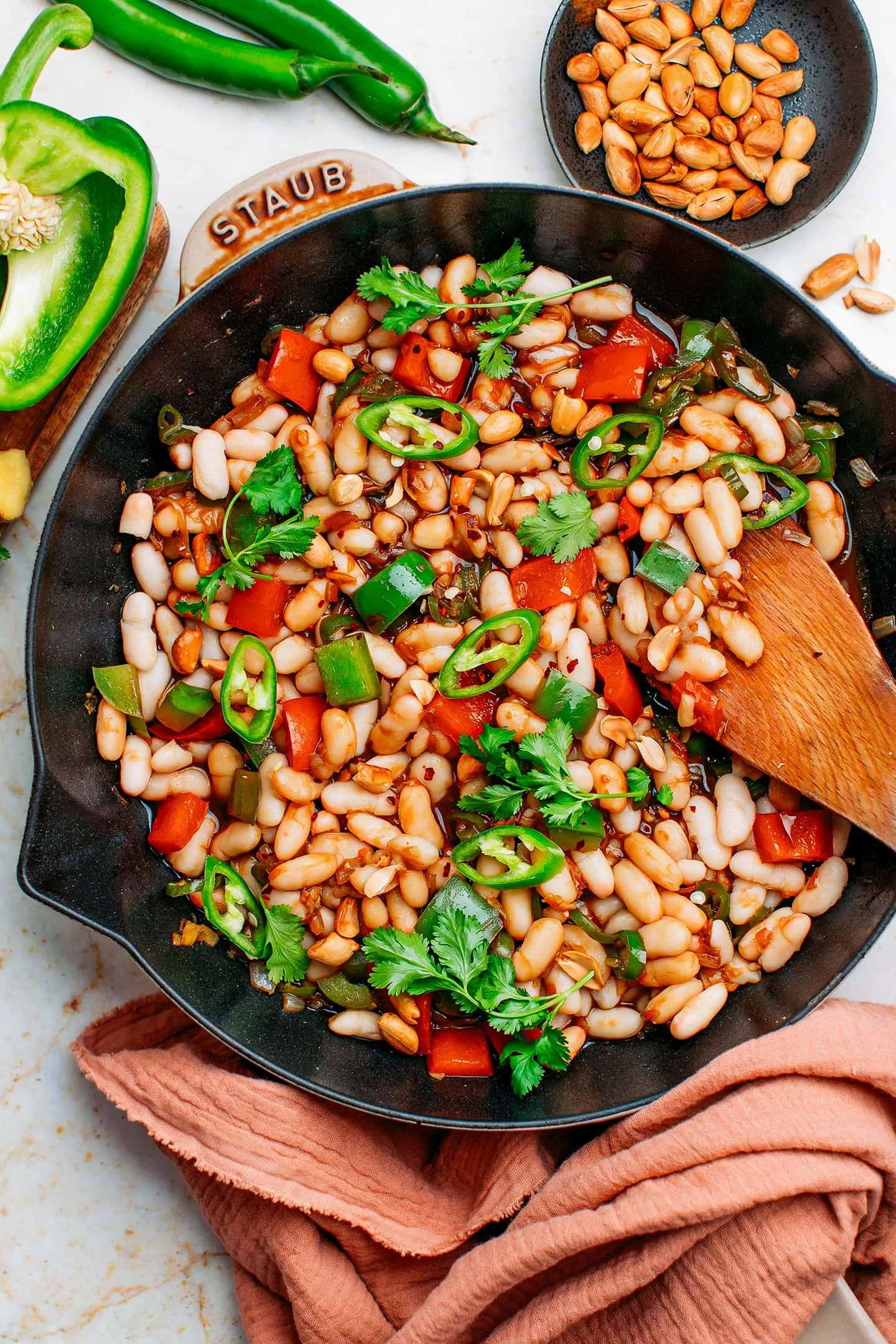Vegetable and bean stir-fry in a black skillet, garnished with cilantro and sliced jalapeños, with a side bowl of peeled garlic cloves.