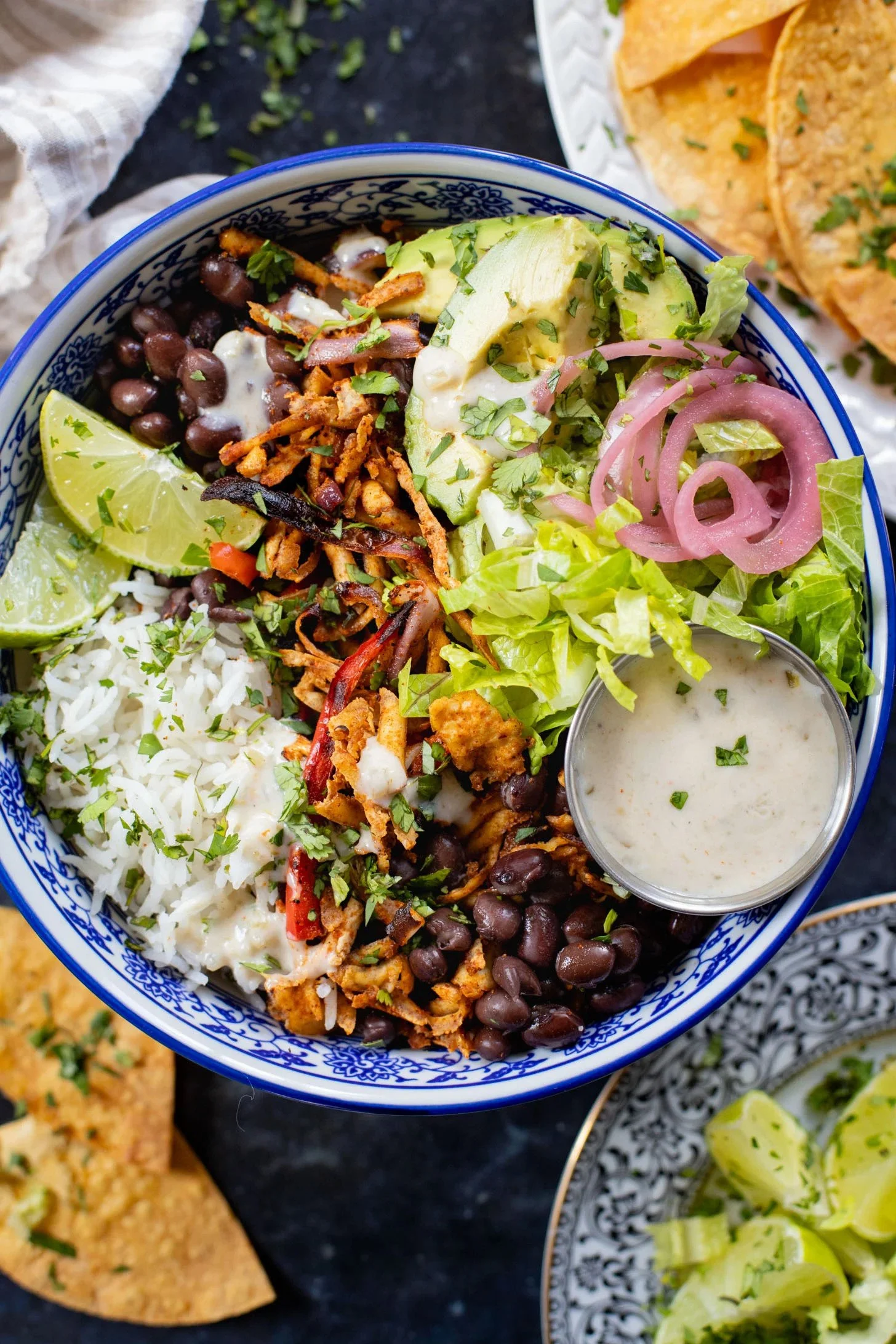 A bowl of Mexican food with rice, black beans, shredded meat, sliced avocado, pickled red onions, lime wedges, lettuce, and a side of creamy dressing.