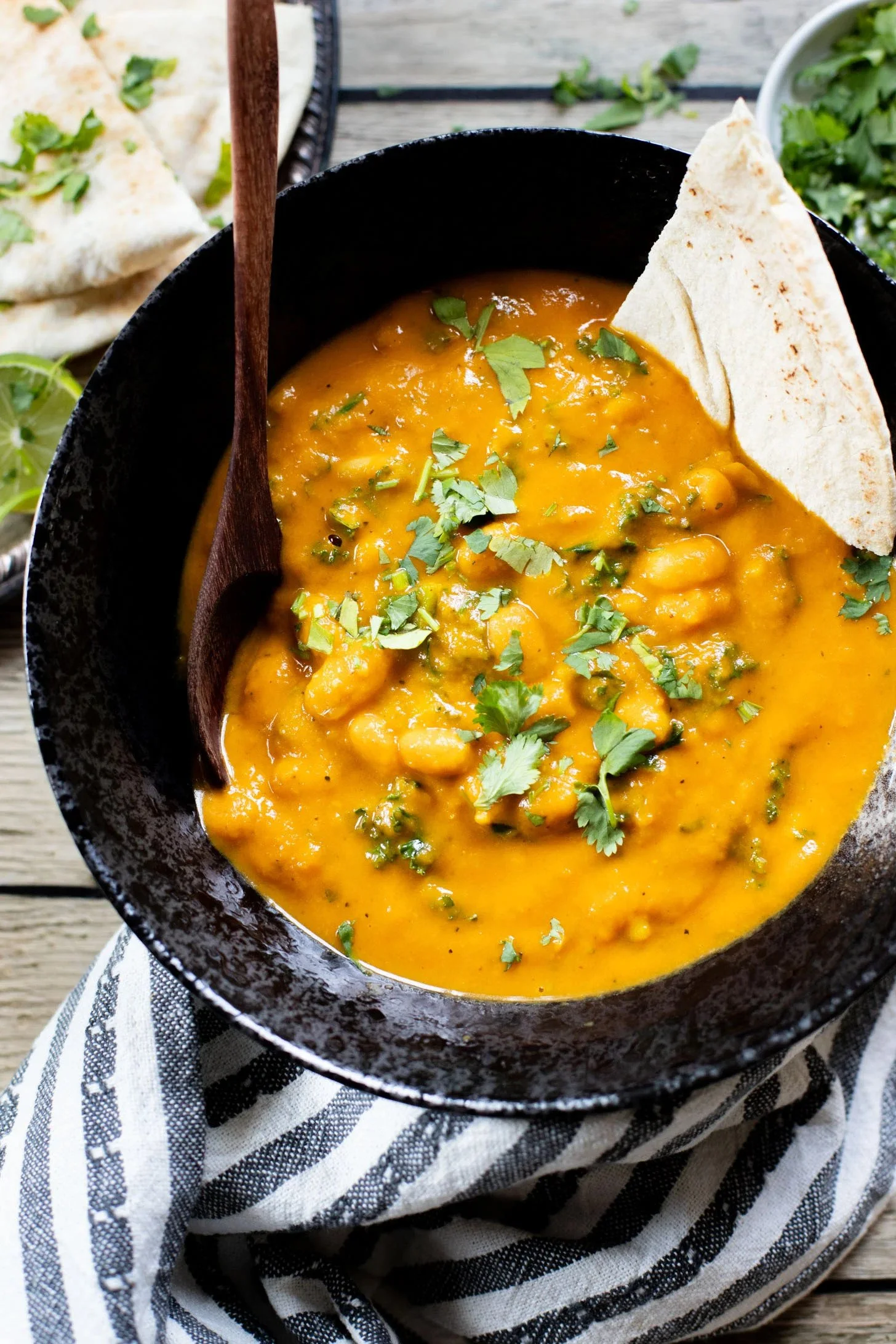 A bowl of yellow chili with cilantro garnish and a piece of flatbread on the side, placed on a black and white striped cloth on a wooden surface.