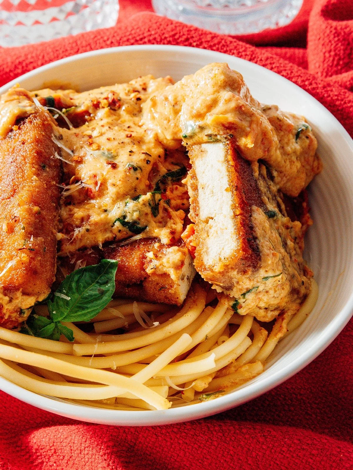 Close-up of a bowl of spaghetti topped with breaded tofu cutlets and creamy sauce, garnished with fresh basil, on a red cloth.