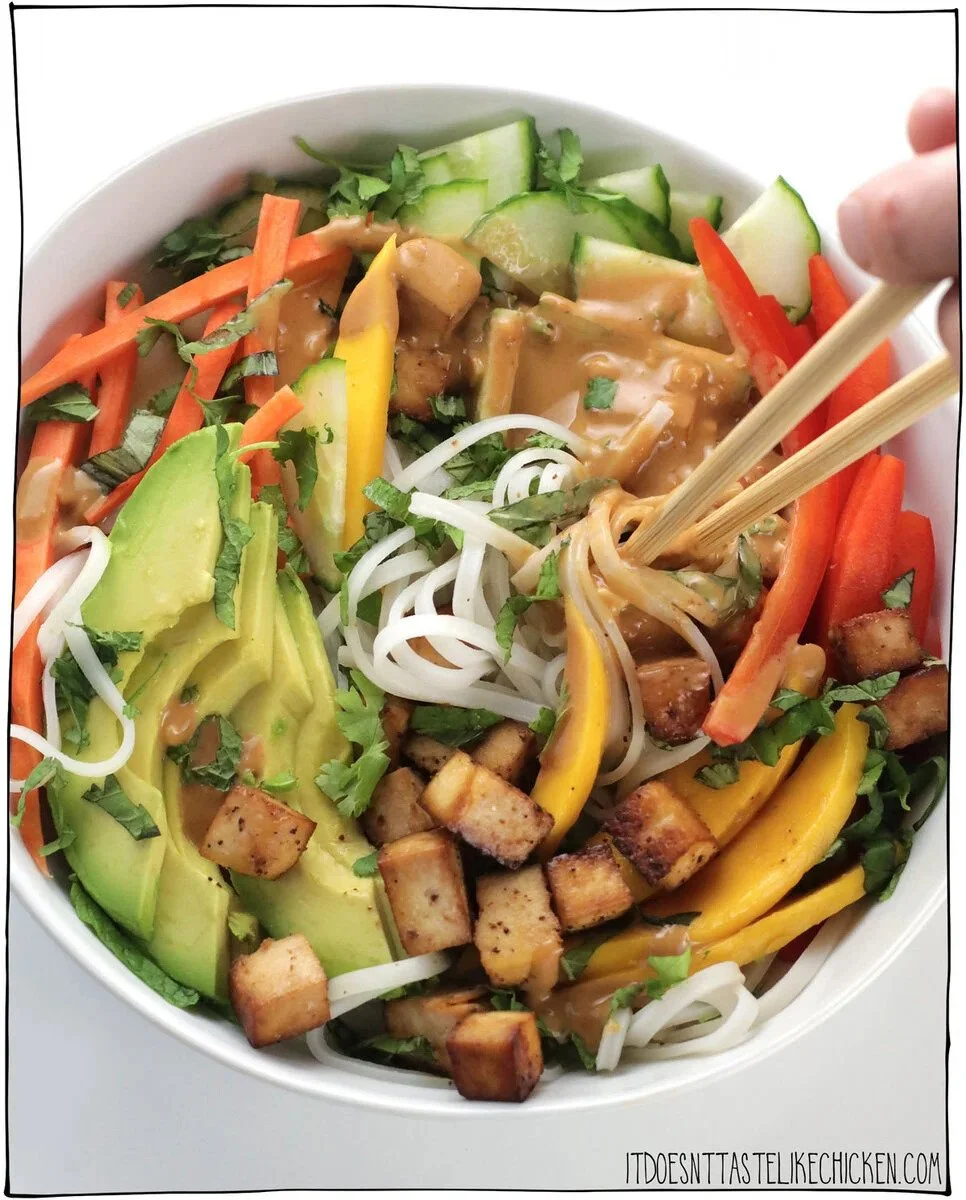 A bowl of vegetable stir-fry containing sliced cucumber, carrots, avocado, red and yellow bell peppers, bean sprouts, chopped cilantro, and pieces of tofu, with chopsticks picking at the food.