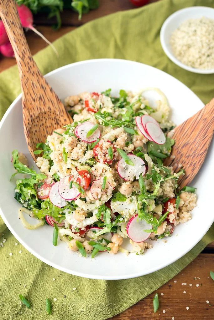 A bowl of couscous salad with radish slices, cherry tomatoes, herbs, and leafy greens, garnished with chopped spring onions.