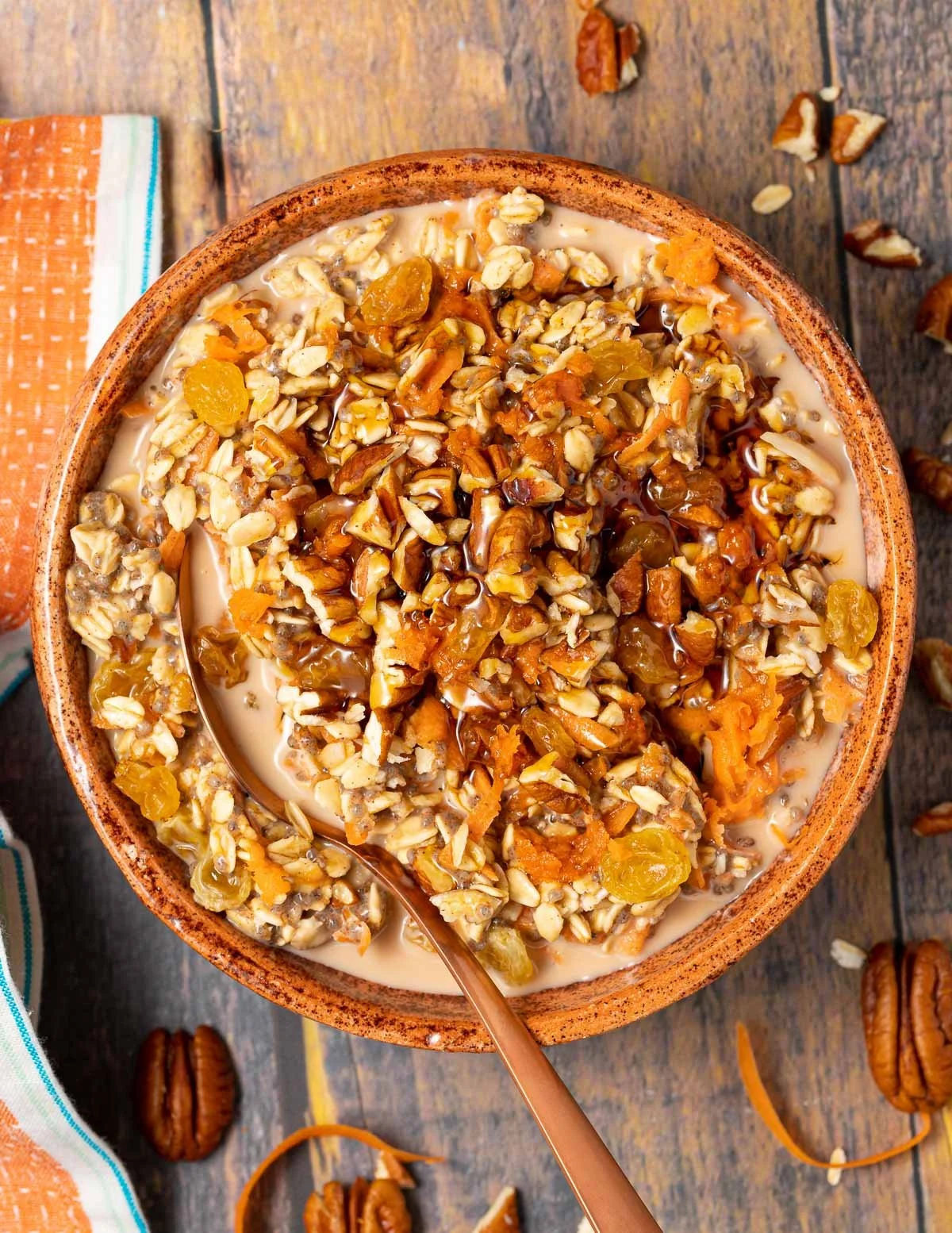 A bowl of oatmeal topped with chopped nuts and dried fruits, served on a rustic wooden surface with scattered pecans and a striped cloth napkin nearby.