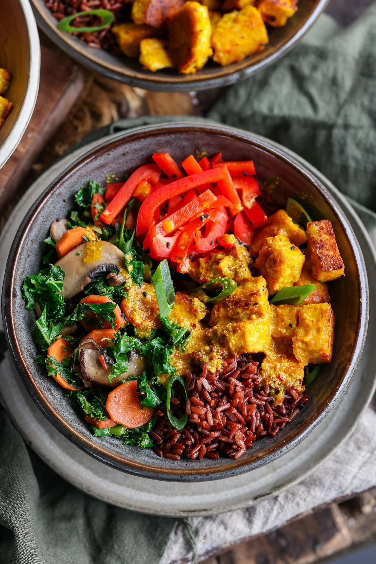 A bowl of vegan Buddha bowl containing red rice, roasted tofu, sautéed kale and mushrooms, sliced carrots, and red bell peppers.