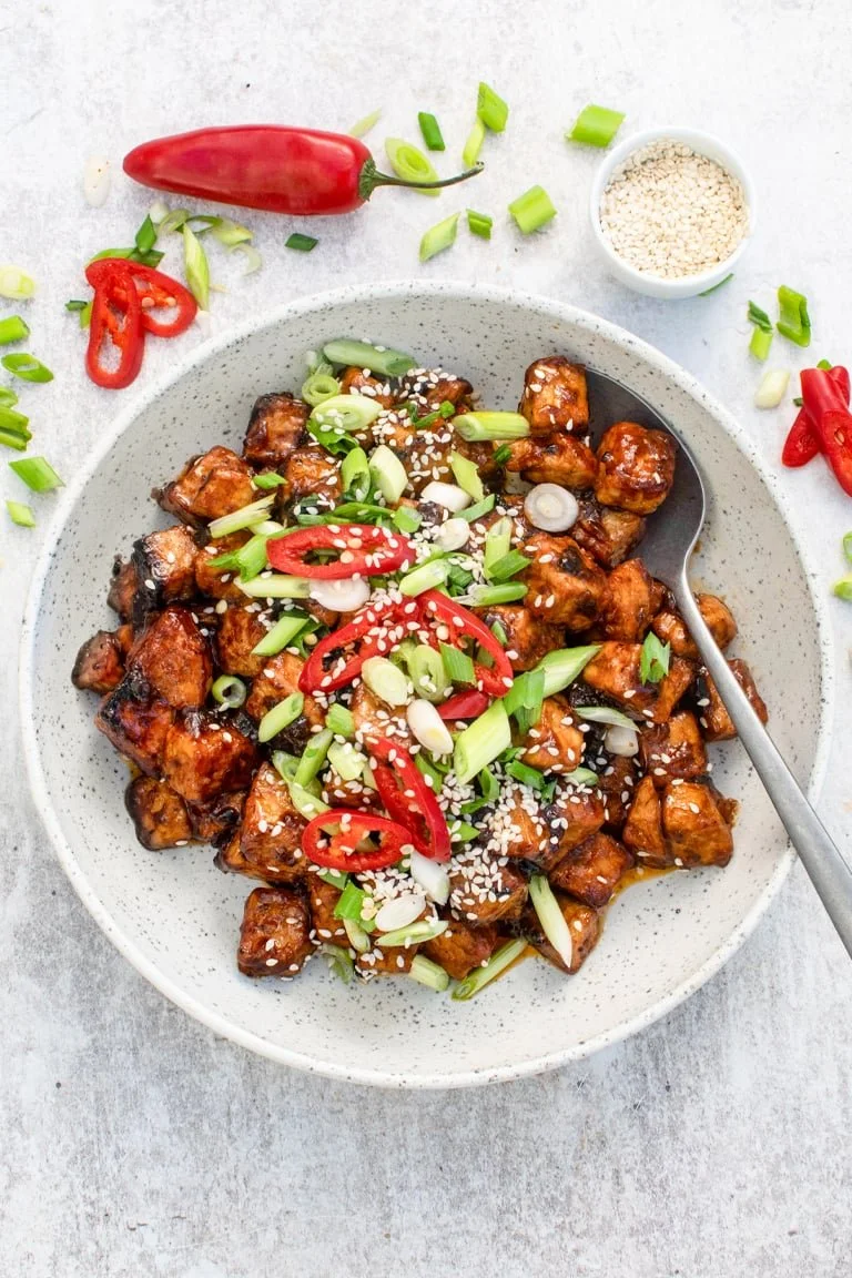 A bowl of glazed tofu bites garnished with sliced green onions, red chili peppers, and sesame seeds, with additional chopped green onions, a red chili pepper, and a small bowl of sesame seeds on a light background