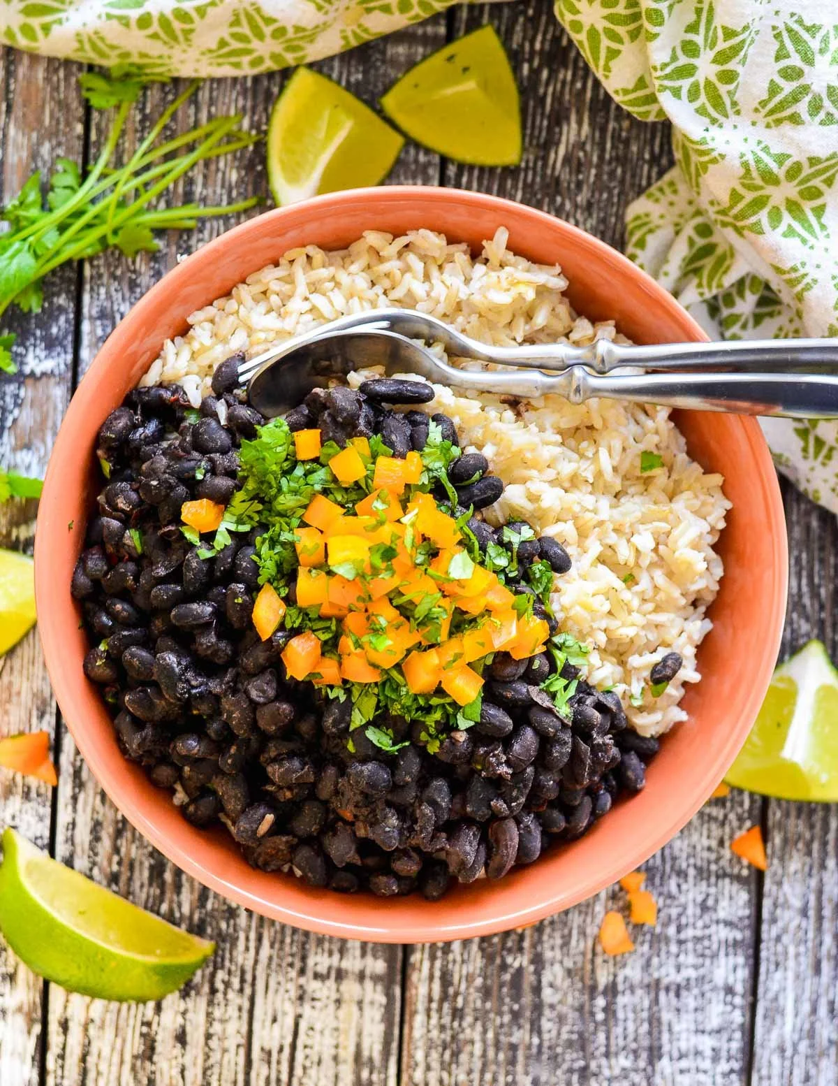A bowl of rice and black beans topped with diced yellow bell peppers and chopped cilantro, garnished with lime wedges and served on a rustic wooden table.