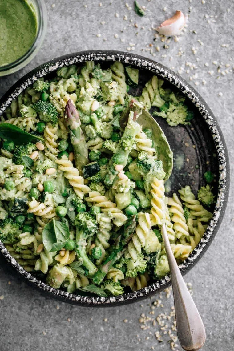Bowl of creamy green vegetable pasta salad made with spiral pasta, broccoli, peas, spinach, and zucchini, with a spoon resting inside and a jar of green sauce nearby on a gray surface with scattered sesame seeds and garlic.