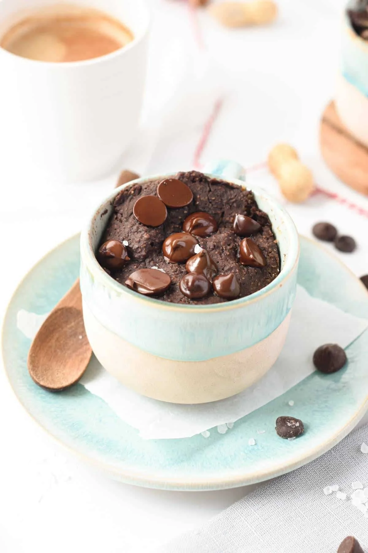 A ceramic bowl of chocolate lava cake topped with melted chocolate chips, placed on a matching plate with a wooden spoon nearby, on a white table with a coffee cup and scattered coffee beans in the background.