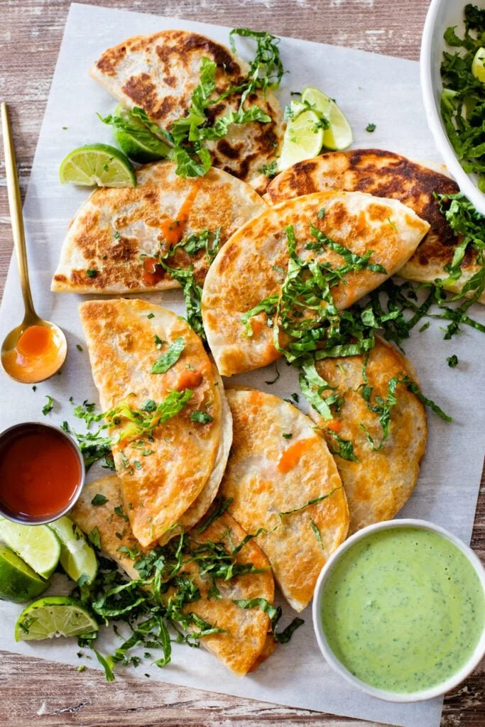 Assorted quesadillas cut into triangles, garnished with chopped cilantro and lime wedges, served with green salsa, hot sauce, and additional lime slices on a wooden table.