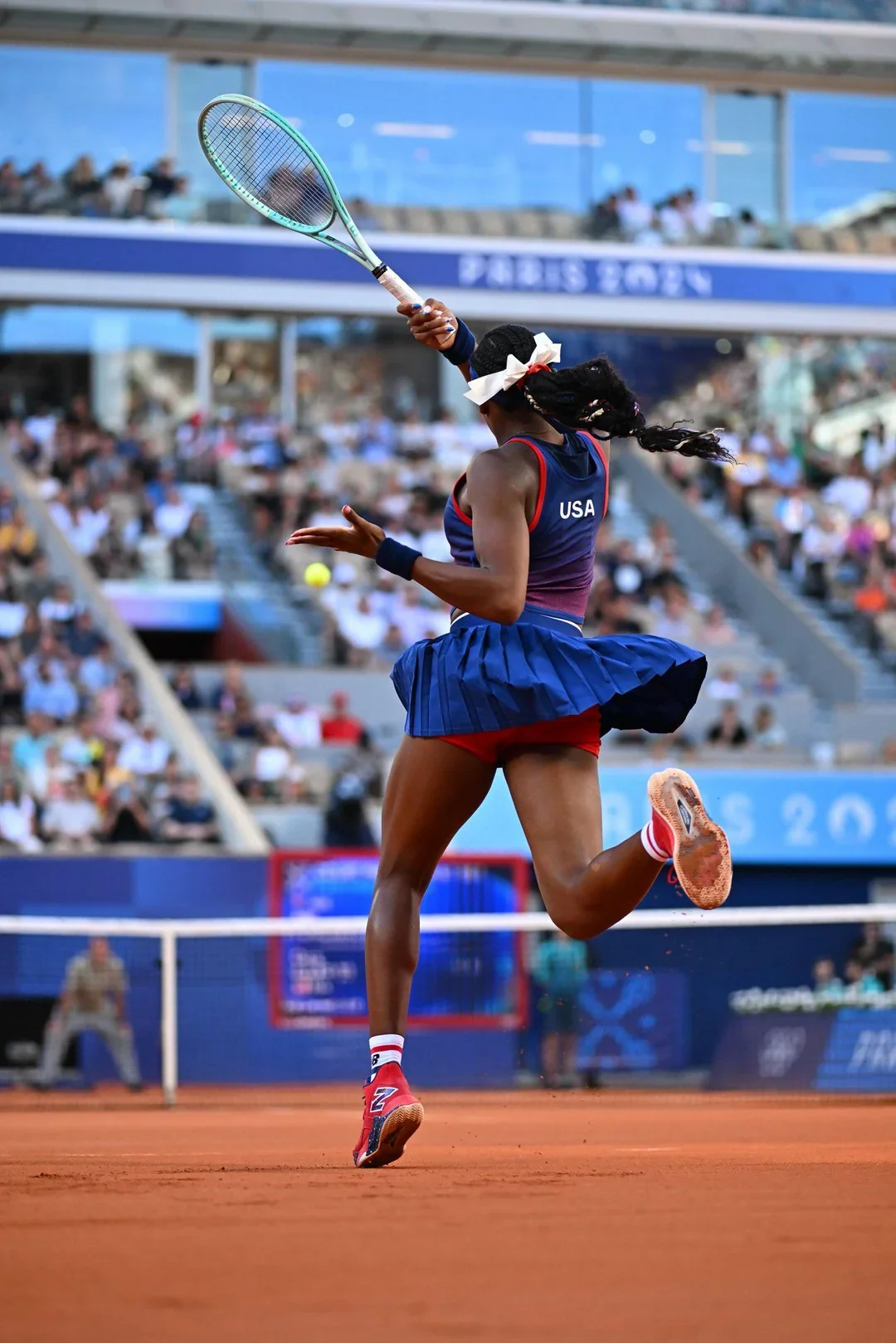 A female athlete wearing a USA tennis uniform is jumping on a clay tennis court while hitting a ball with a tennis racket during a match at the Olympics.
