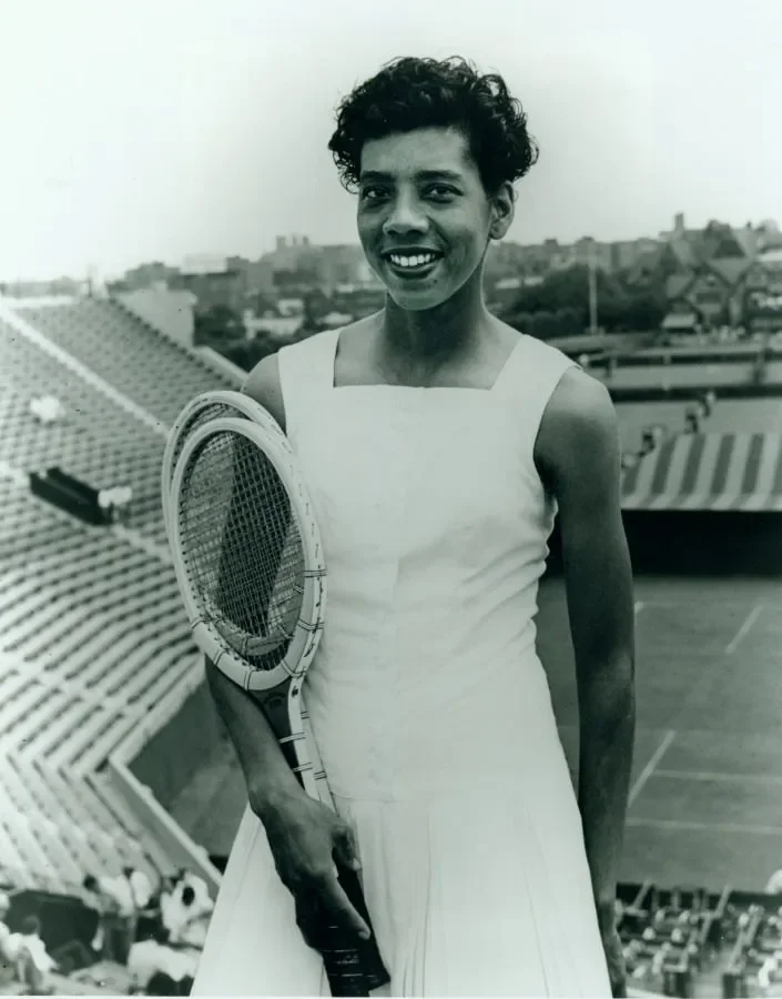 A woman in a white dress holding a tennis racket, smiling, at a stadium tennis court.