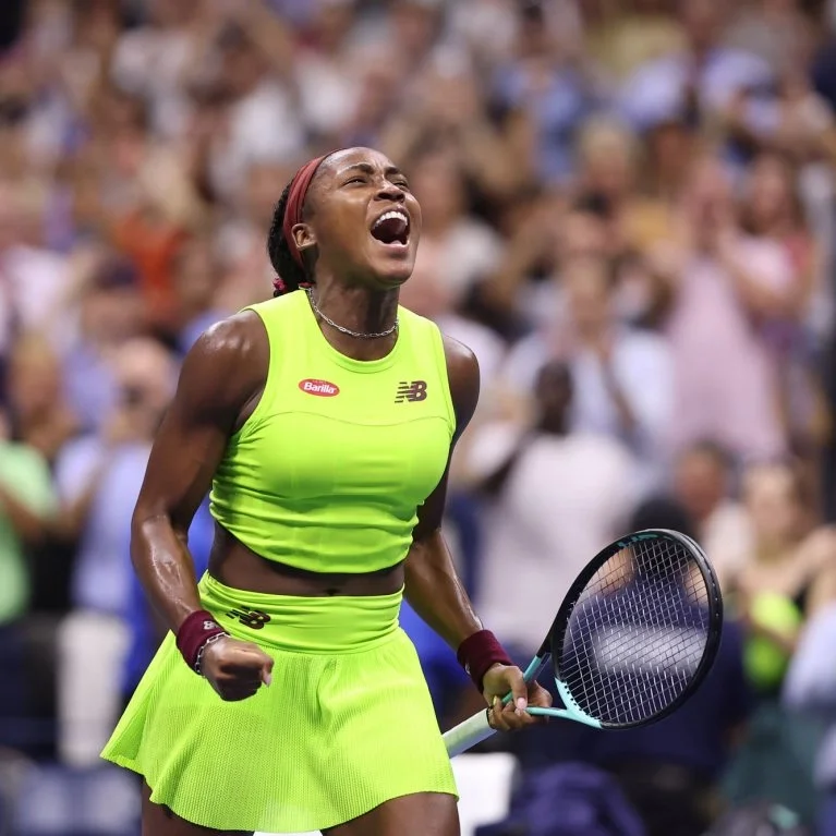 Female tennis player in a neon yellow outfit celebrating on the court with a tennis racket in hand, with a crowd in the background.