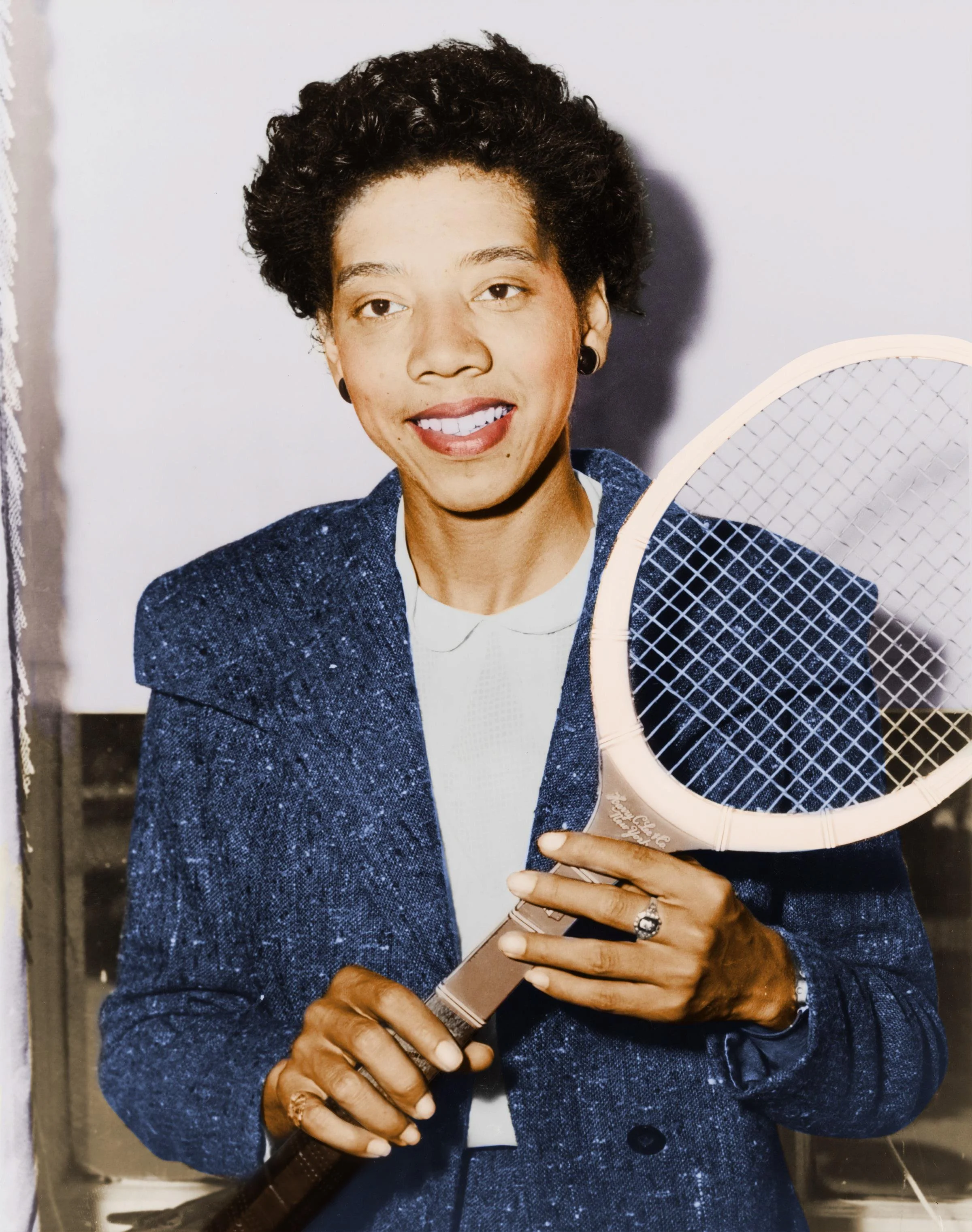 A woman with short curly hair smiling and holding a tennis racket, dressed in a blue suit and white shirt.