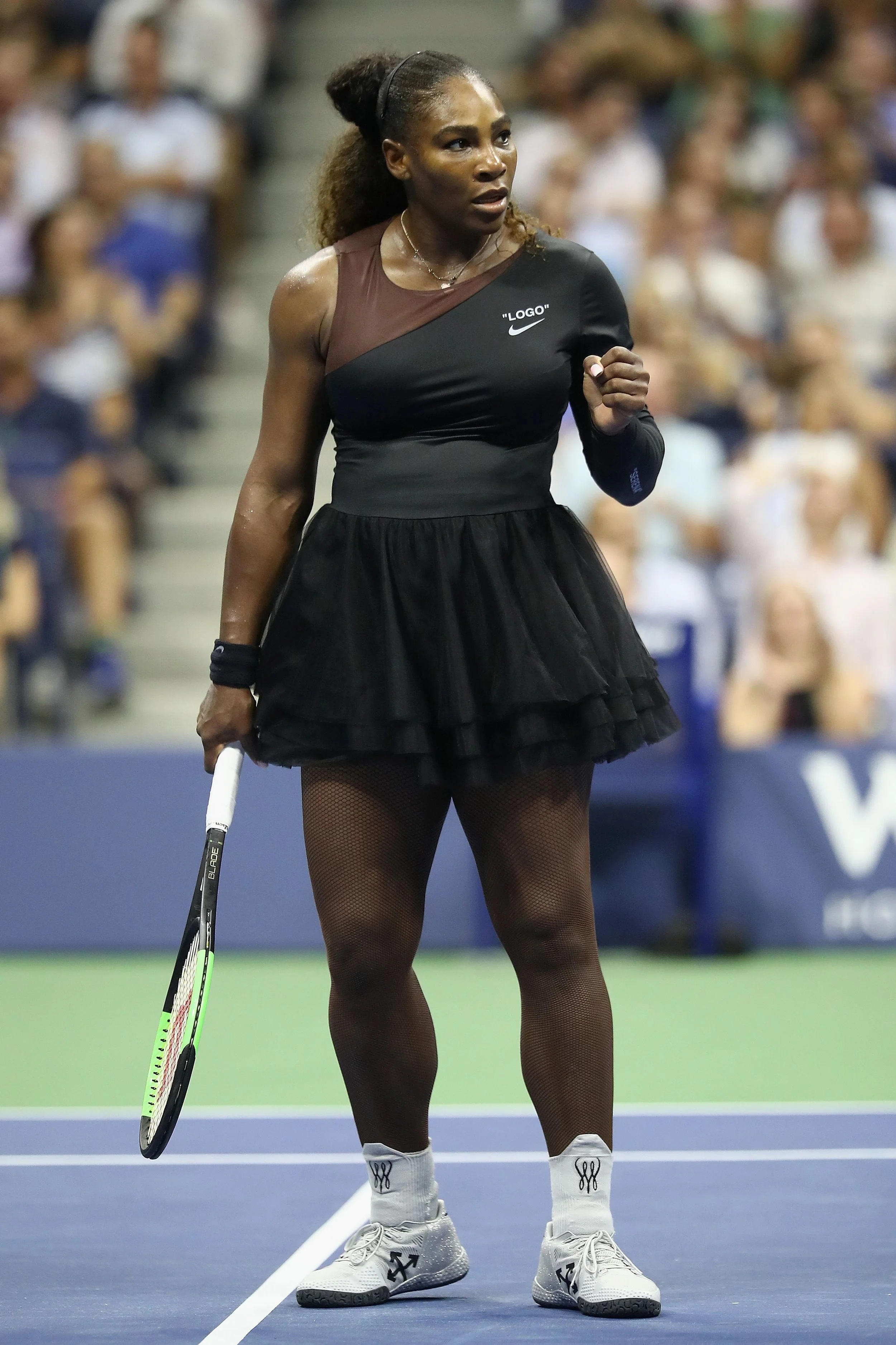 A female tennis player standing on a tennis court holding a racket, wearing a black tutu-like skirt, white sneakers, black fishnet stockings, and a black and brown sleeveless top, with a crowd in the background.